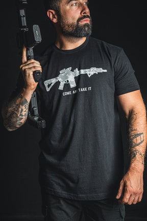 A bearded man holds a rifle over his shoulder while wearing the Born United Private Label “Come and Take It” tee, which features a bold graphic of a rifle and the phrase COME AND TAKE IT printed on the front—a striking statement in support of 2nd Amendment rights.