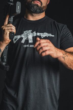A man holding a firearm wears the Born United Private Label “Come and Take It” tee—a black patriotic shirt featuring a rifle graphic and the statement “COME AND TAKE IT”—captured against a dark background as a bold tribute to the 2nd Amendment.