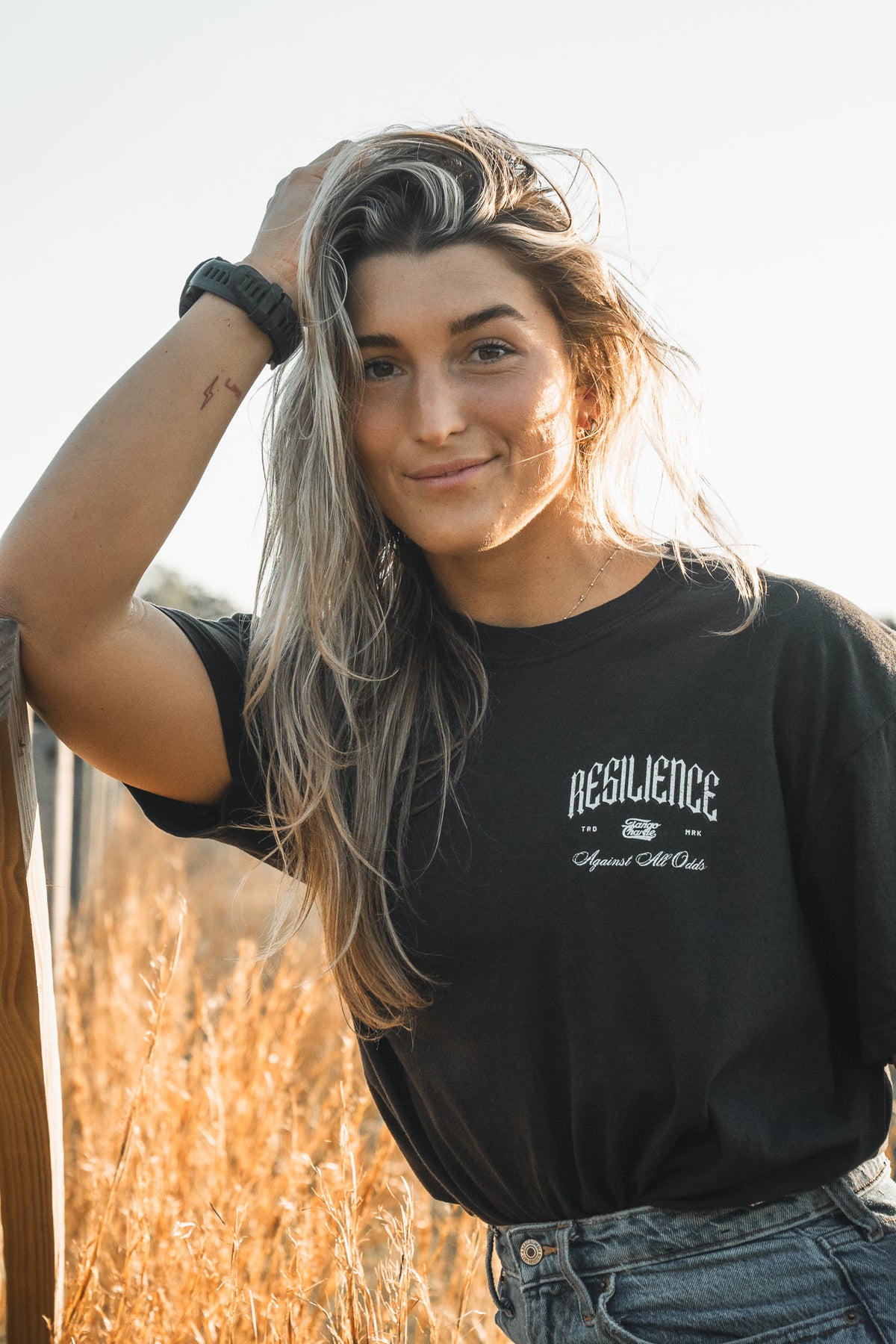 A woman with long, wavy hair stands in a field wearing the Resilience - Premium Tee by Tango Charlie Apparel and jeans, smiling softly with one hand on her head and her arm resting on a wooden fence.