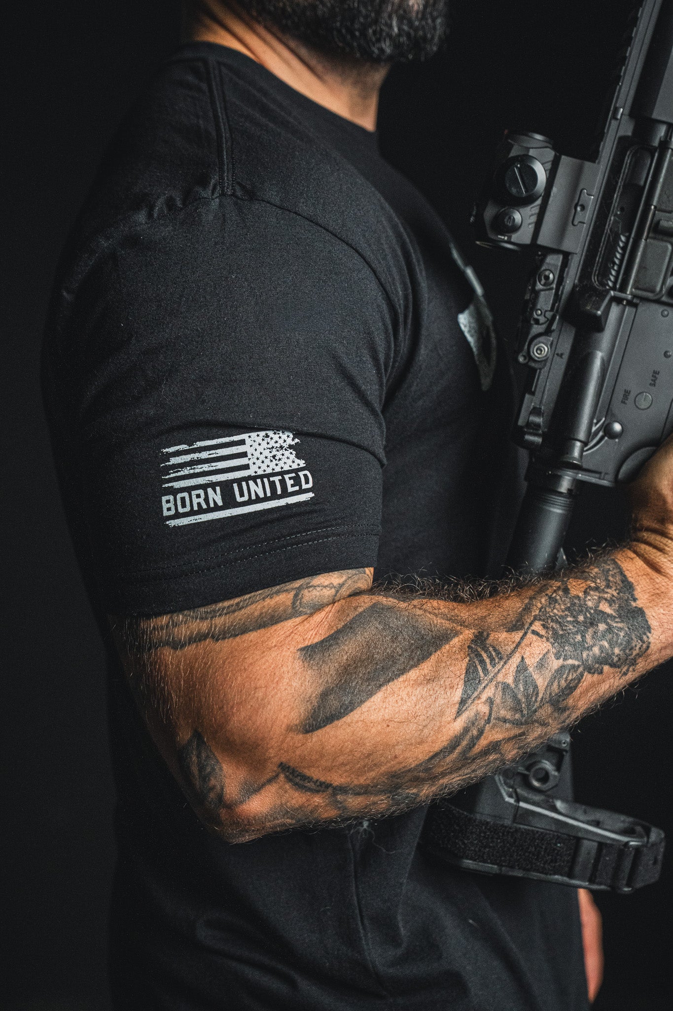 A man with a tattooed arm holds a rifle, wearing the Born United Private Label “Come and Take It” black patriotic tee, which features a stylized American flag and the words BORN UNITED on the sleeve—a bold statement celebrating the 2nd Amendment.