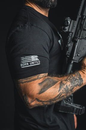 A man with a tattooed arm holds a rifle, wearing the Born United Private Label “Come and Take It” black patriotic tee, which features a stylized American flag and the words BORN UNITED on the sleeve—a bold statement celebrating the 2nd Amendment.