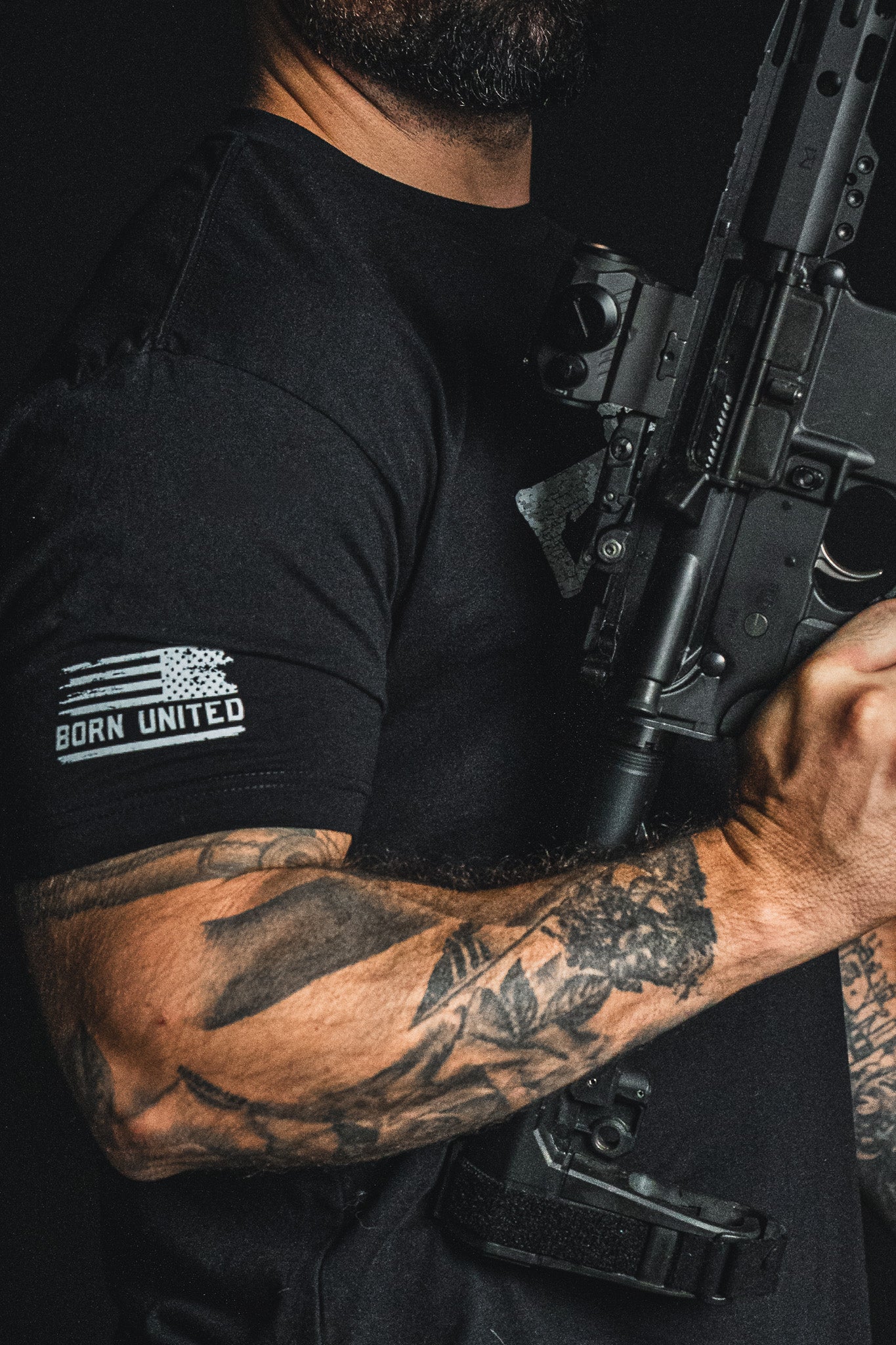 A tattooed person wears the Born United Private Label “Come and Take It” black patriotic tee, featuring “BORN UNITED” lettering, while holding a black rifle; only the upper body and part of the firearm are visible.