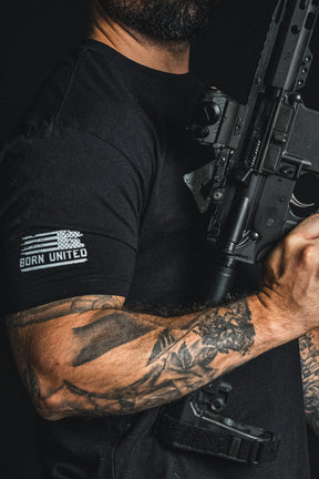 A tattooed person wears the Born United Private Label “Come and Take It” black patriotic tee, featuring “BORN UNITED” lettering, while holding a black rifle; only the upper body and part of the firearm are visible.