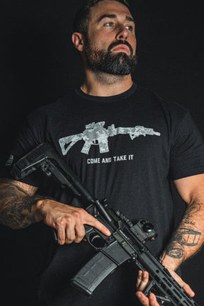 A tattooed man holds a black rifle and wears the Born United Private Label “Come and Take It” tee, which features a bold rifle graphic and the statement text “COME AND TAKE IT” against a dark background, making a strong 2nd Amendment statement.