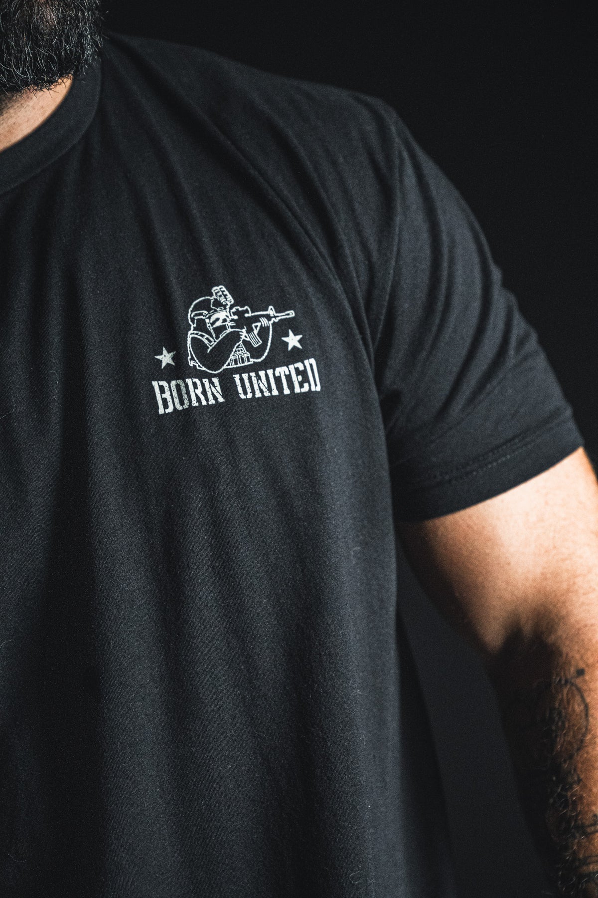 A person wearing a black “Tactically Delayed” cotton t-shirt from Born United Private Label, featuring a white graphic of a soldier holding a rifle and “BORN UNITED” printed on the chest.