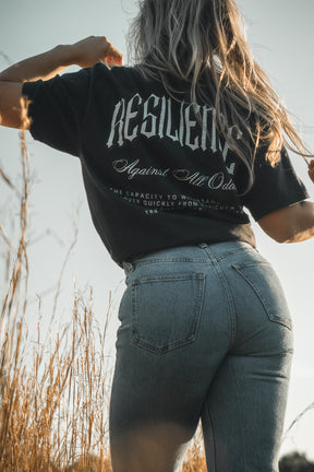 Basking in sunlight in a field of tall grass, they wear light blue jeans and the Tango Charlie Apparel "Resilience - Premium Tee": a breathable, comfy black shirt boldly featuring RESILIENCE on the back.