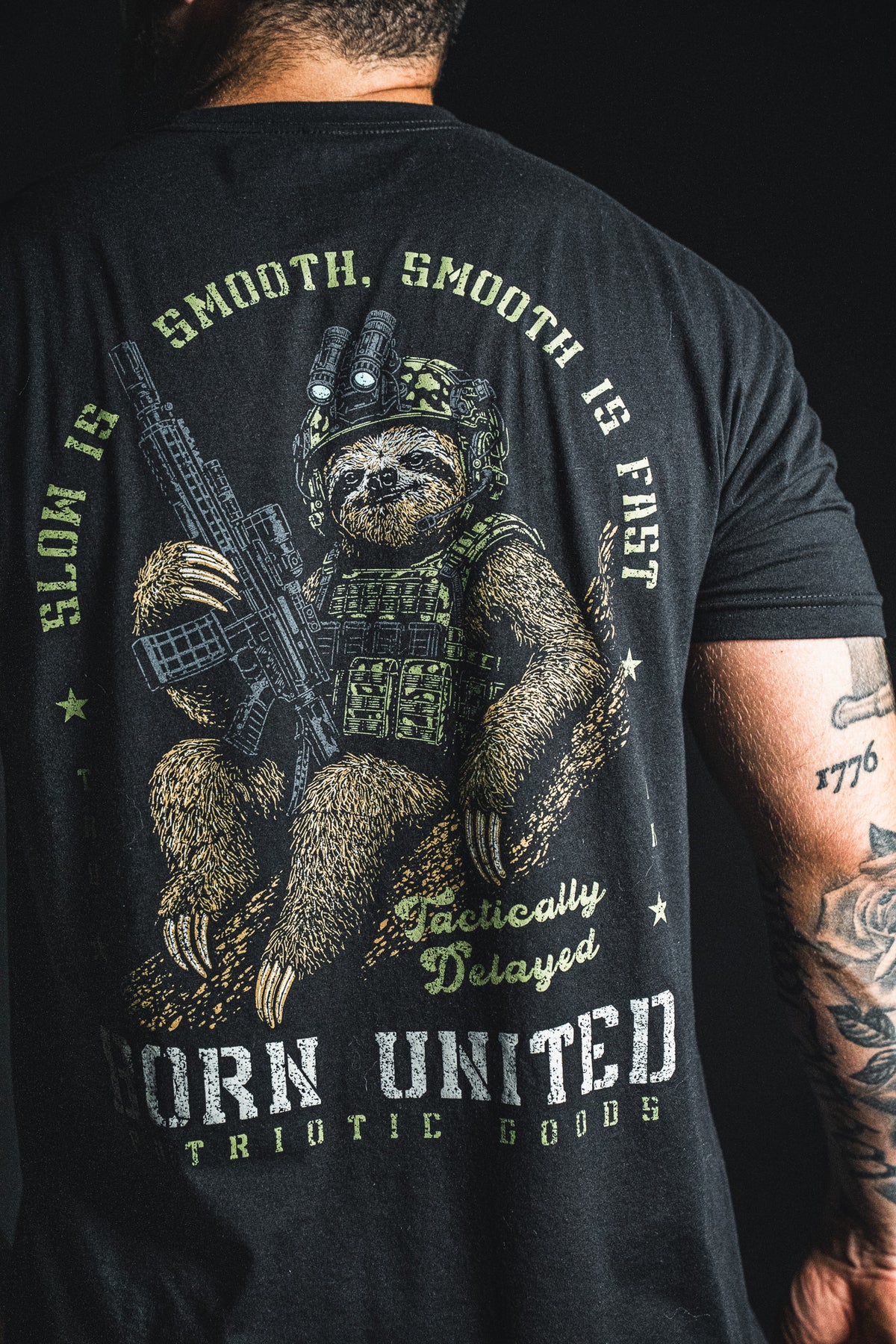 A person wearing the Born United Private Label “Tactically Delayed” shirt, a black cotton tee featuring an illustrated tactical sloth equipped with a rifle, helmet, and vest, along with the phrases “Slow is smooth, smooth is fast” and “Tactically delayed.” This humorous shirt is perfect for anyone who enjoys clever humor and a relaxed style.