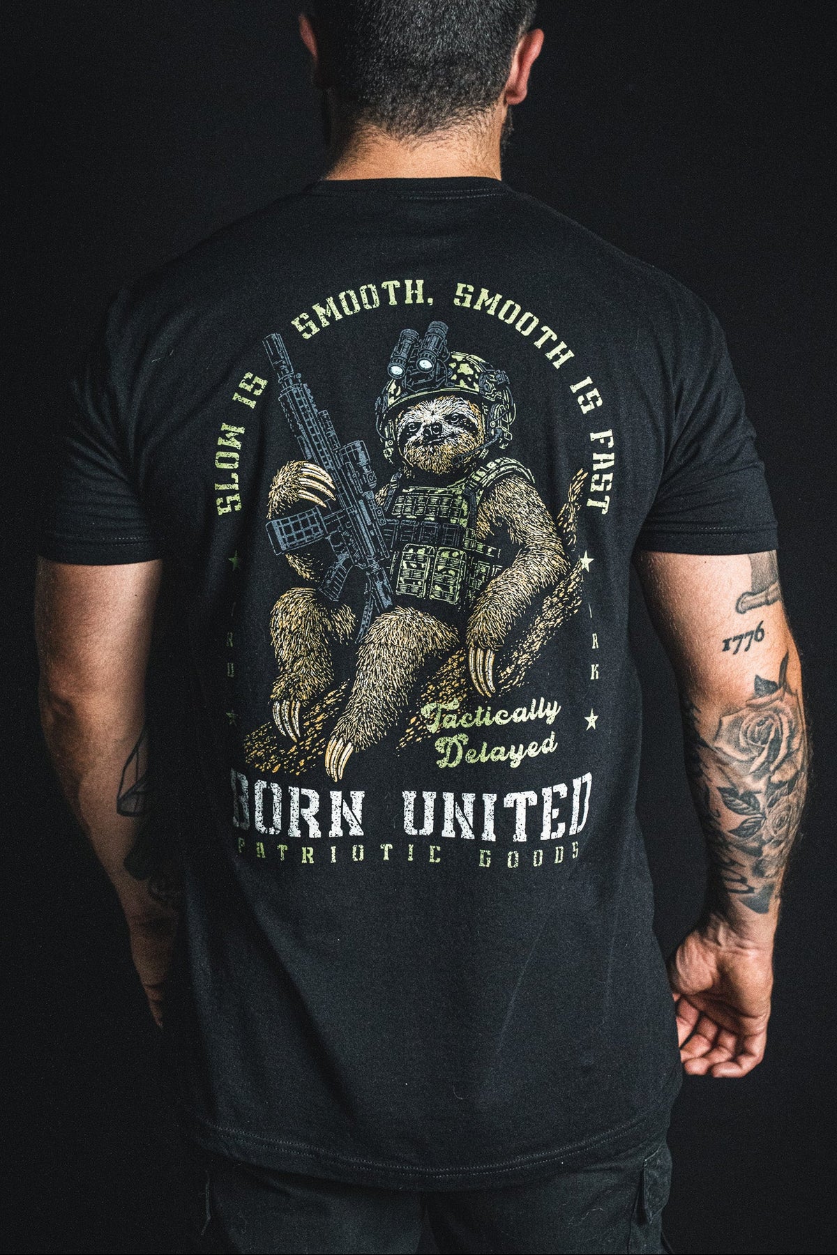 A tattooed man faces away, wearing the Tactically Delayed tee by Born United Private Label—this black shirt displays an armed sloth in tactical gear alongside the phrases “Slow is smooth, smooth is fast” and “Born United Patriotic Goods,” adding humor to any outfit.