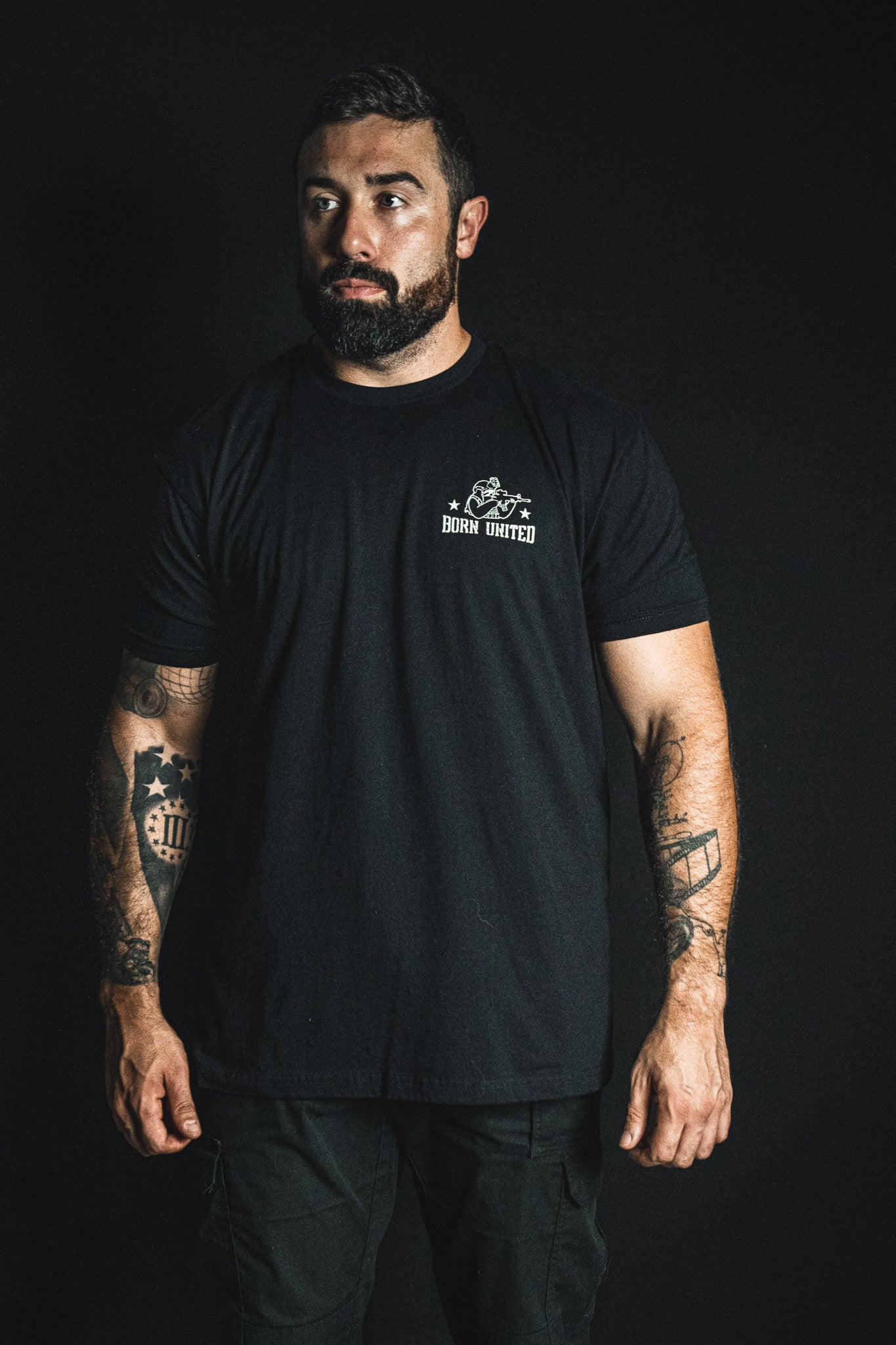 A bearded man wears a black cotton Tactically Delayed Tee with a small Born United graphic of a rifleman on the left chest.