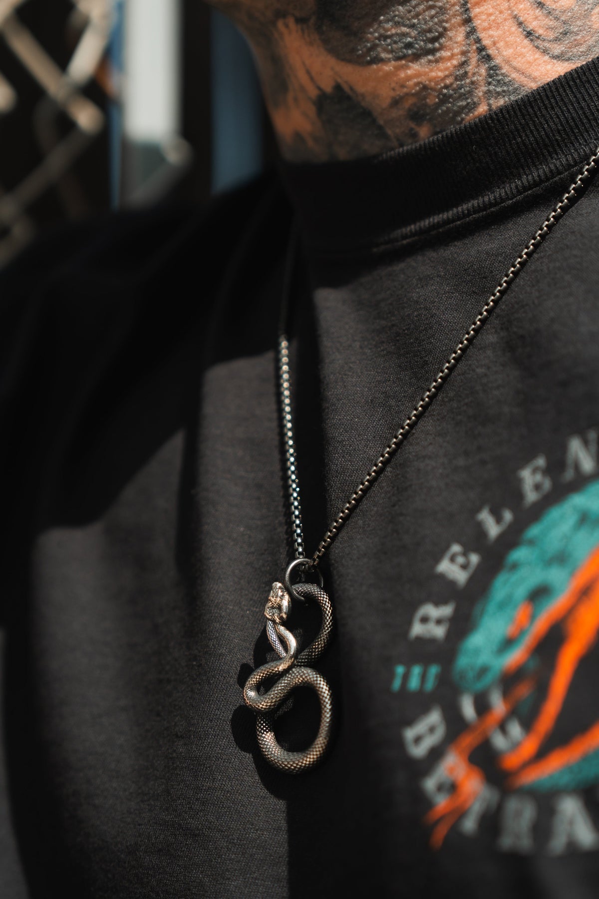 Close-up of a person wearing a black shirt with colorful graphics, showcasing serpentine elegance with the Slither Necklace by Relentless Betrayal. The background is slightly blurred, revealing neck tattoos, as the statement piece features an intricate snake pendant.