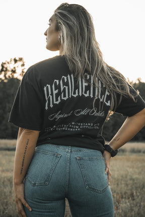 A woman in a field, facing away, wears the Tango Charlie Apparel "Resilience - Premium Tee" with "RESILIENCE Against All Odds" and blue jeans. Her hair is loose, tattoos visible, surrounded by trees and grass.