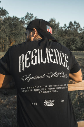A person stands by a wooden fence outdoors in a Tango Charlie Apparel "Resilience - Premium Tee" and a black cap with a US flag patch, surrounded by trees.