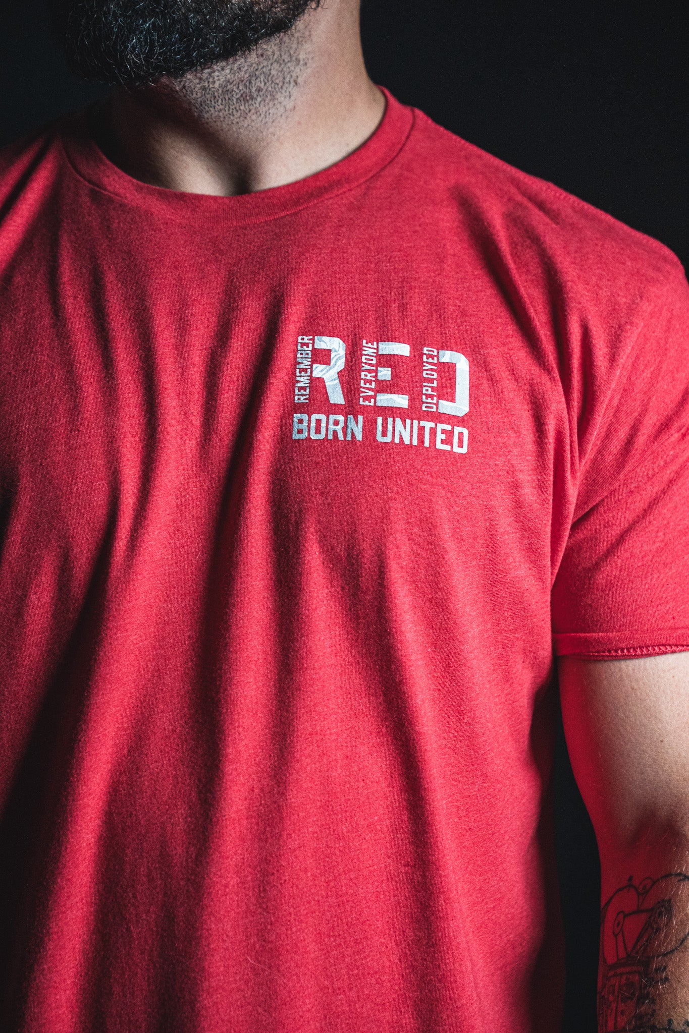 A person wearing a Born United Private Label RFV2 red T-shirt, featuring the text RED BORN UNITED printed on the upper chest. Only the torso, right arm, and part of the face are visible, with the shirt’s vibrant color subtly conveying a Remember Everyone Deployed message.