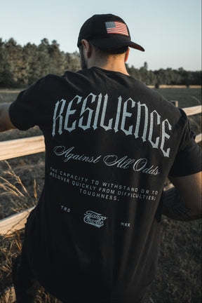 A man in a Tango Charlie Apparel black Resilience - Premium Tee and a black cap with an American flag patch leans on a wooden fence outdoors, surrounded by trees and tall grass.