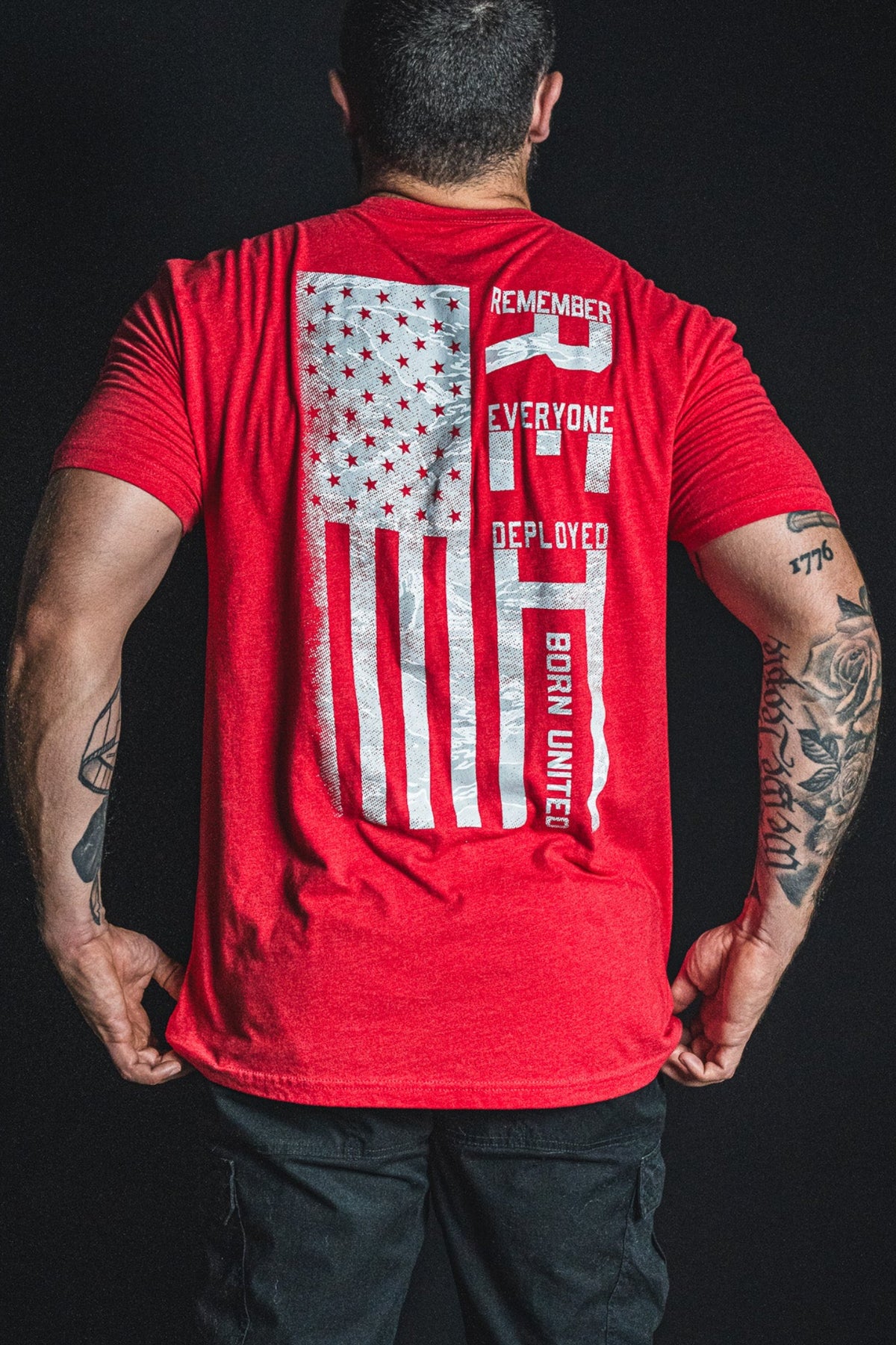 A man faces away, wearing the Born United Private Label RFV2 red shirt featuring a distressed American flag and the phrases “REMEMBER EVERYONE DEPLOYED” and “BORN UNITED.” His arms display various tattoos.