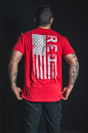 A man faces away, wearing the Born United Private Label RFV2 red shirt featuring a distressed American flag and the phrases “REMEMBER EVERYONE DEPLOYED” and “BORN UNITED.” His arms display various tattoos.