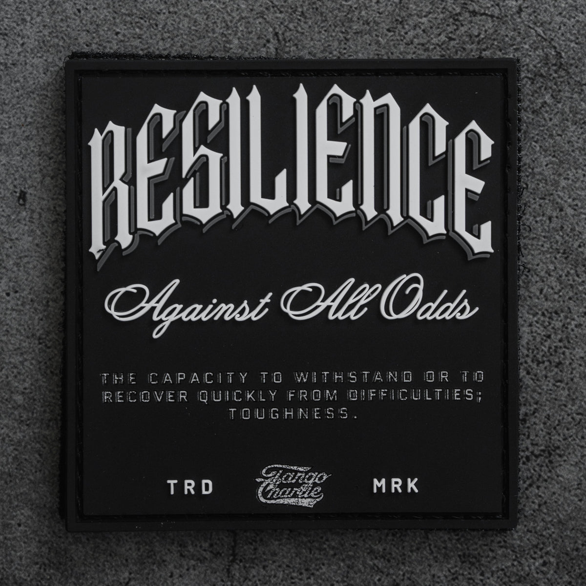 The Resilience - Patch by Tango Charlie Apparel is a black 3-inch Velcro patch with bold white lettering: “RESILIENCE Against All Odds,” shown here on a textured gray surface.