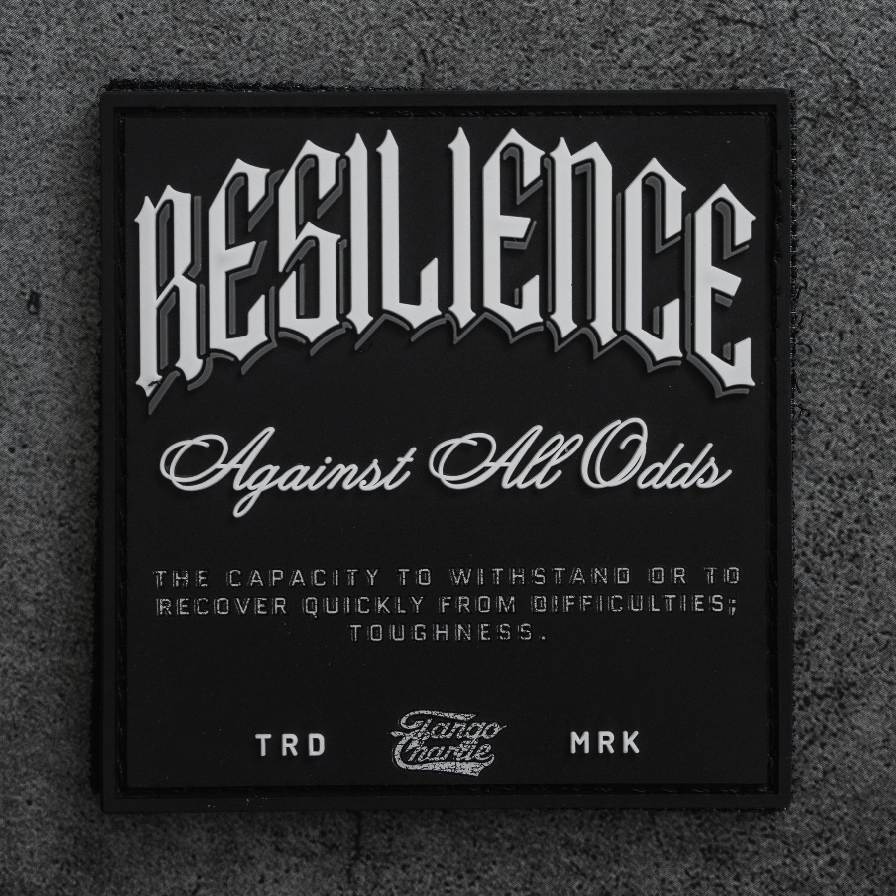 The Resilience - Patch by Tango Charlie Apparel is a black 3-inch Velcro patch with bold white lettering: “RESILIENCE Against All Odds,” shown here on a textured gray surface.