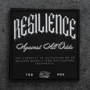 The Resilience - Patch by Tango Charlie Apparel is a black 3-inch Velcro patch with bold white lettering: “RESILIENCE Against All Odds,” shown here on a textured gray surface.