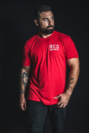 A man with tattoos wears a red Born United Private Label RFV2 T-shirt and black pants, standing against a dark background—a subtle nod to the “Remember Everyone Deployed” message.
