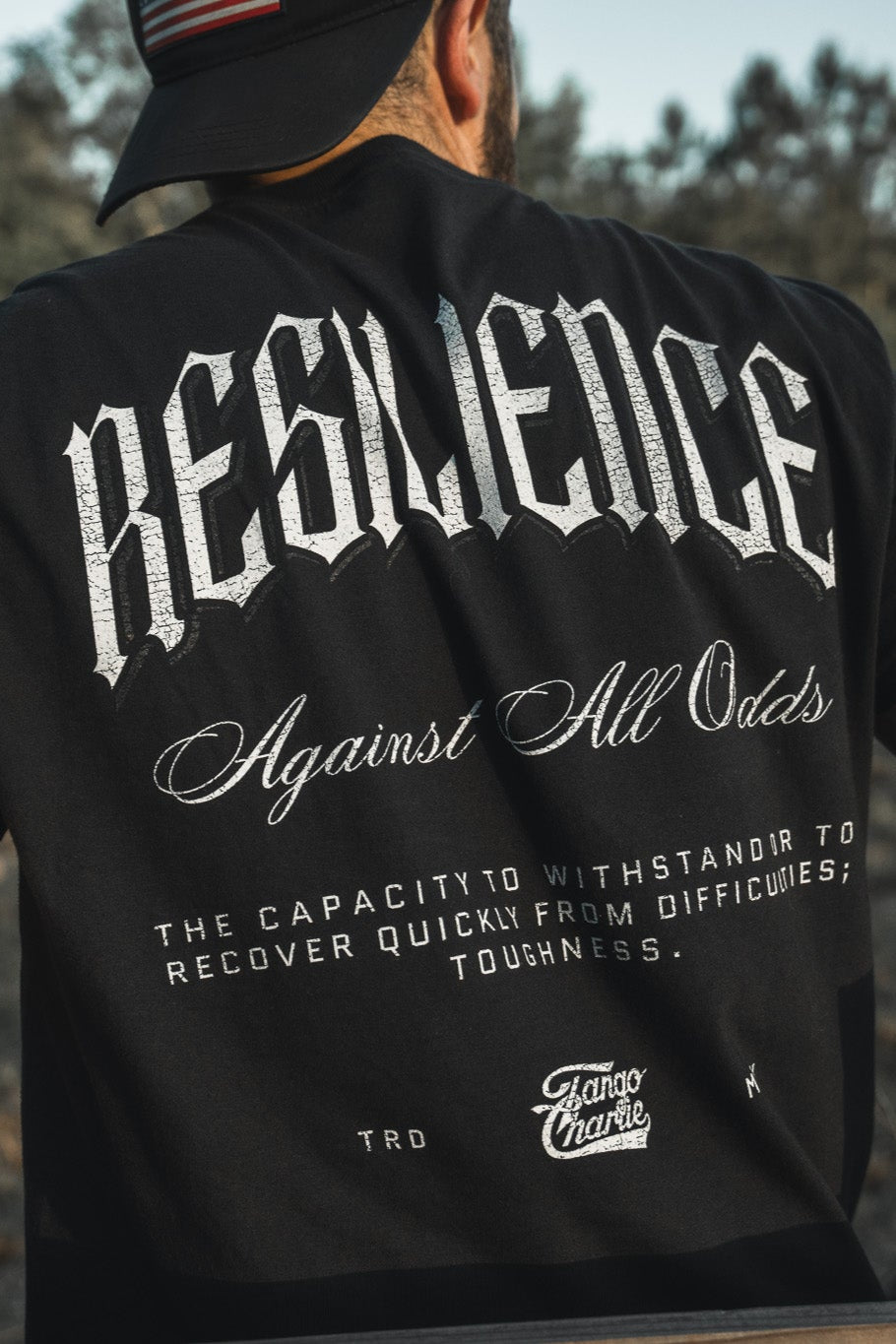 A person wears the Tango Charlie Apparel Resilience - Premium Tee, a black heavyweight hemp shirt with bold white "RESILIENCE Against All Odds" text on the back—tough, breathable comfort that stands up to challenges.