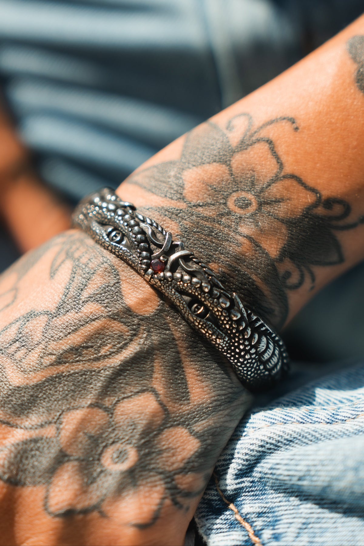 A tattooed arm, adorned with floral designs, rests on denim fabric, wearing the Holy Grail Bracelet by Relentless Betrayal. The textured, dark metallic piece is shaped like a dragon with intricate details, evoking a mythical artifact against the softly focused background.