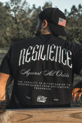 A man wears the Resilience - Premium Tee by Tango Charlie Apparel—a black, breathable shirt and cap with a US flag patch. The tee reads: “RESILIENCE Against All Odds. The capacity to withstand or to recover quickly from difficulties; toughness.”.