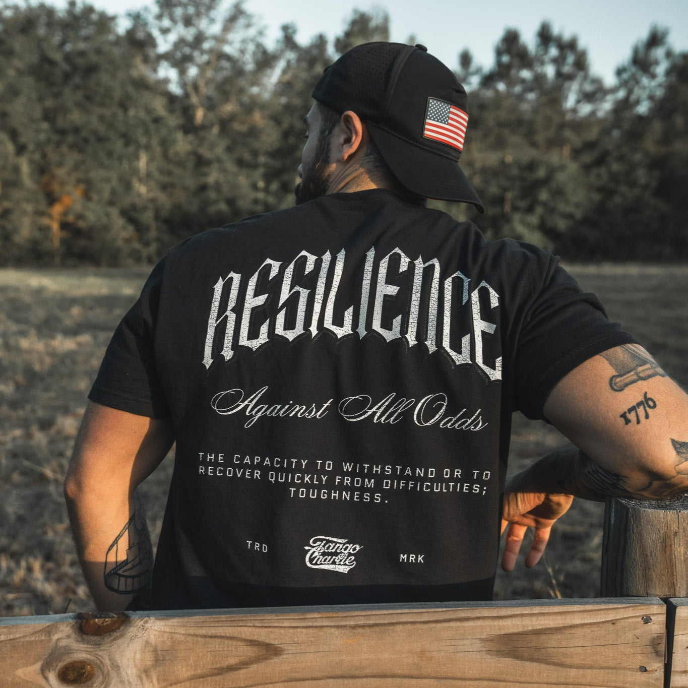 A man in a Tango Charlie Apparel Resilience - Premium Tee and a black cap with an American flag patch sits on a wooden fence, surrounded by trees and grass. The tee features "RESILIENCE Against All Odds" with its definition on the back.