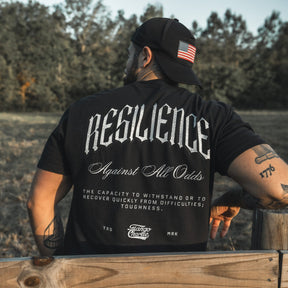 A man in a Tango Charlie Apparel Resilience - Premium Tee and a black cap with an American flag patch sits on a wooden fence, surrounded by trees and grass. The tee features "RESILIENCE Against All Odds" with its definition on the back.