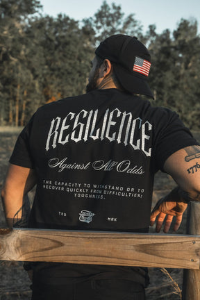 A man in a Tango Charlie Apparel black cap with an American flag patch and a "Resilience - Premium Tee" stands by a wooden fence outdoors, trees visible behind him.