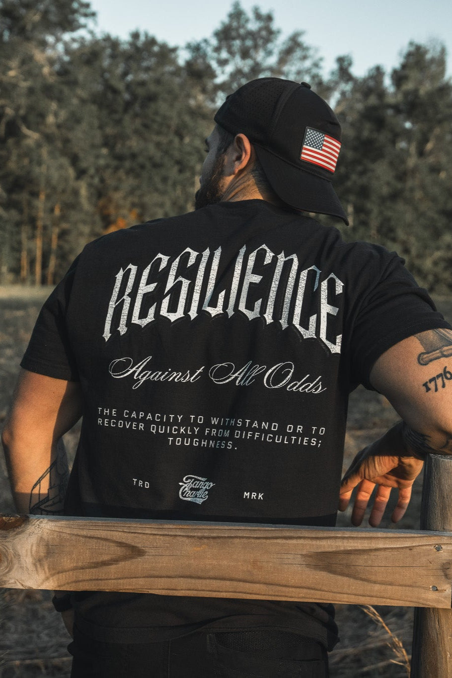 A man in a Tango Charlie Apparel black cap with an American flag patch and a "Resilience - Premium Tee" stands by a wooden fence outdoors, trees visible behind him.