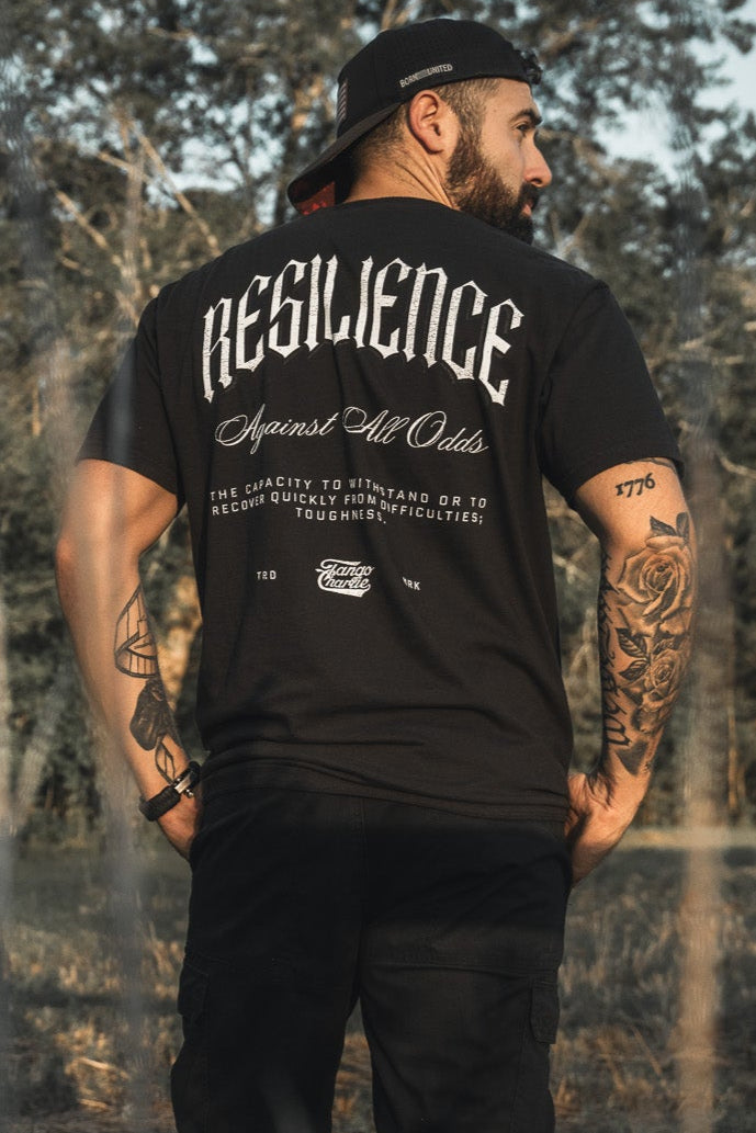 A bearded man stands outdoors with his back to the camera, wearing a black cap and the Tango Charlie Apparel "Resilience - Premium Tee." The shirt reads RESILIENCE Against All Odds. His tattooed arms are visible among trees and sunlight.