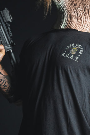 A person wearing the “G.O.A.T.” patriotic statement tee from Born United Private Label holds a firearm, their face partially covered by a mask with an animal skin pattern.