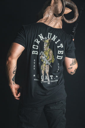 A person wearing a ram mask holds a rifle, displaying the back of the Born United Private Label G.O.A.T. tee, which features a military goat graphic and the text “Born United, Stay True, G.O.A.T., Guardian of American Tradition”—a patriotic shirt honoring American tradition.
