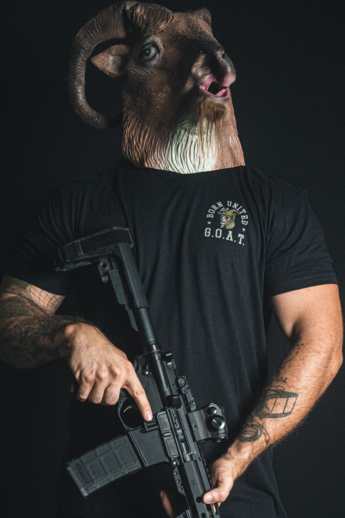 A person in a ram mask and a Born United Private Label G.O.A.T. tee holds a black rifle against a dark background.