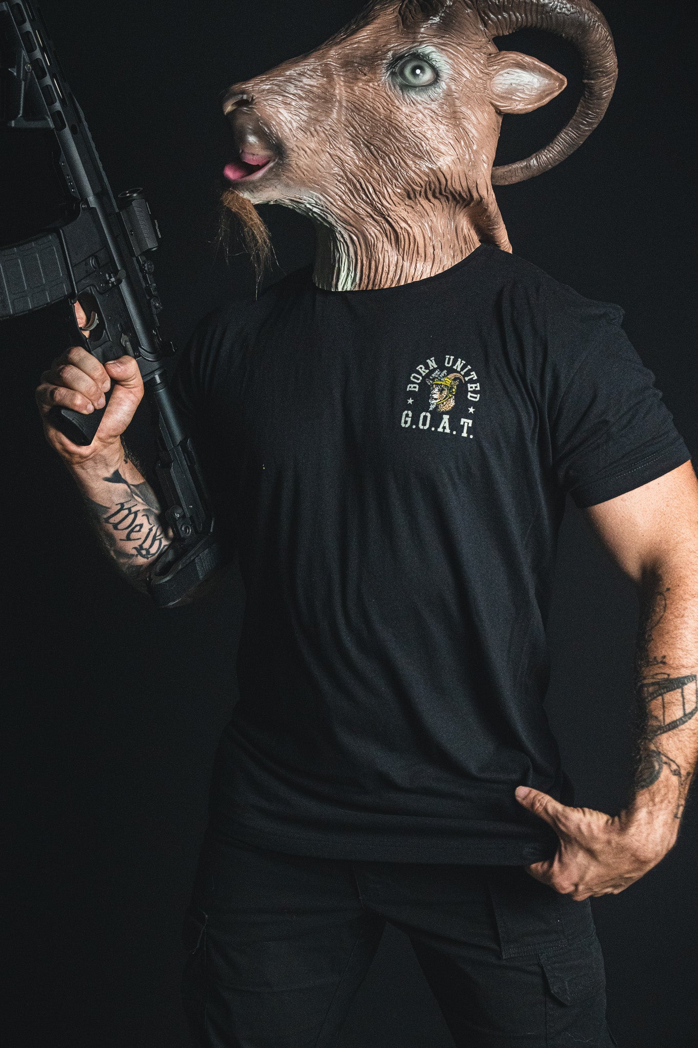 A person wearing a goat mask and the “G.O.A.T.” statement tee from Born United Private Label, holding a rifle with visible arm tattoos, stands against a dark background.