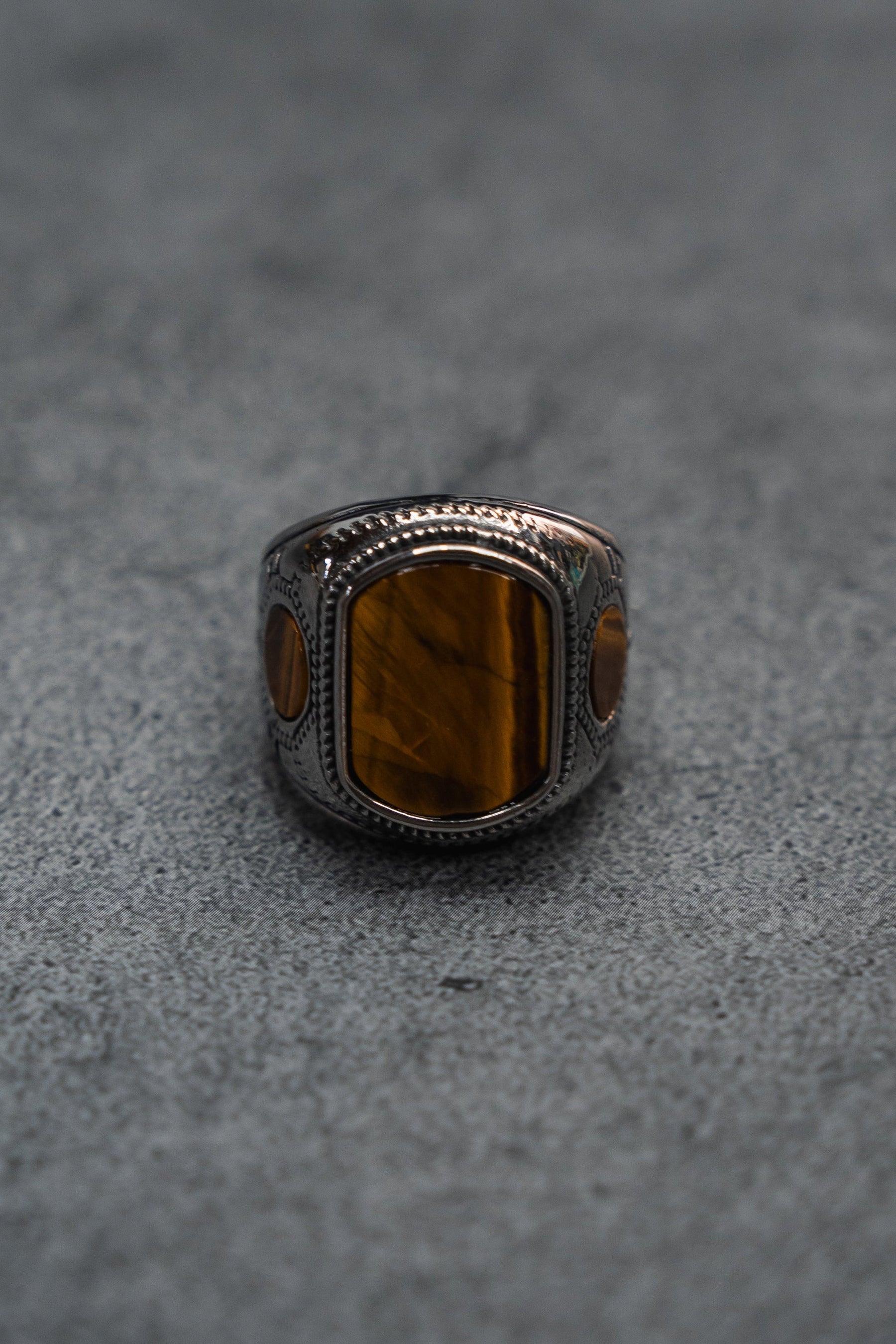 The Desert Relic Ring by Relentless Betrayal is a handcrafted silver band showcasing a rectangular brown stone at its center, beautifully presented on a minimalist gray surface.