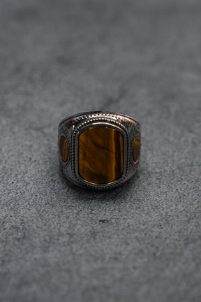 The Desert Relic Ring by Relentless Betrayal is a handcrafted silver band showcasing a rectangular brown stone at its center, beautifully presented on a minimalist gray surface.