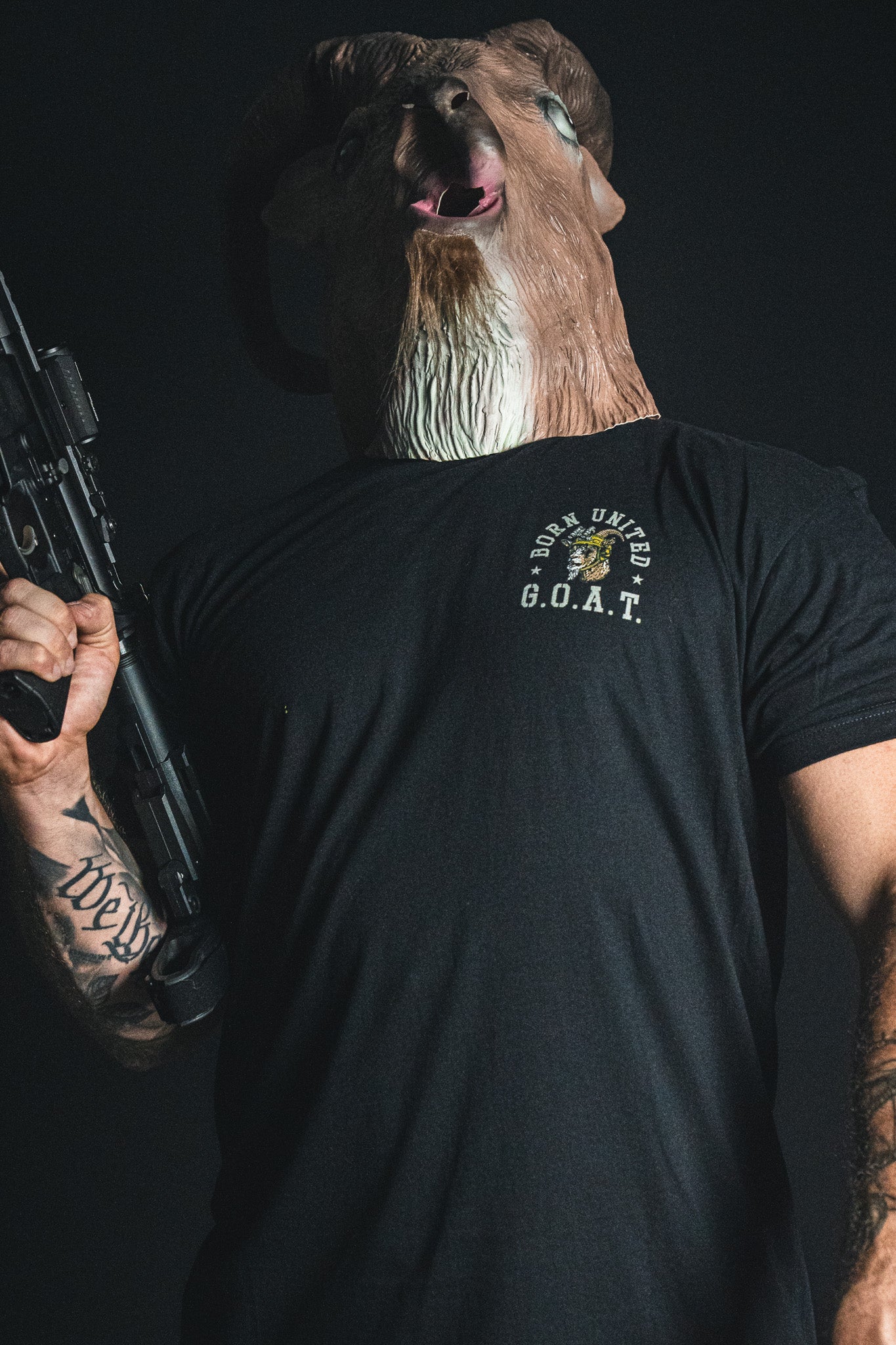 A person wearing a ram mask and holding a rifle stands against a dark background, dressed in the black “G.O.A.T.” statement tee from Born United Private Label.