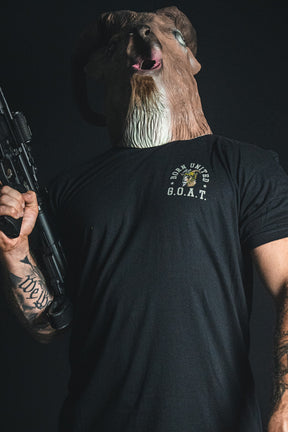A person wearing a ram mask and holding a rifle stands against a dark background, dressed in the black “G.O.A.T.” statement tee from Born United Private Label.