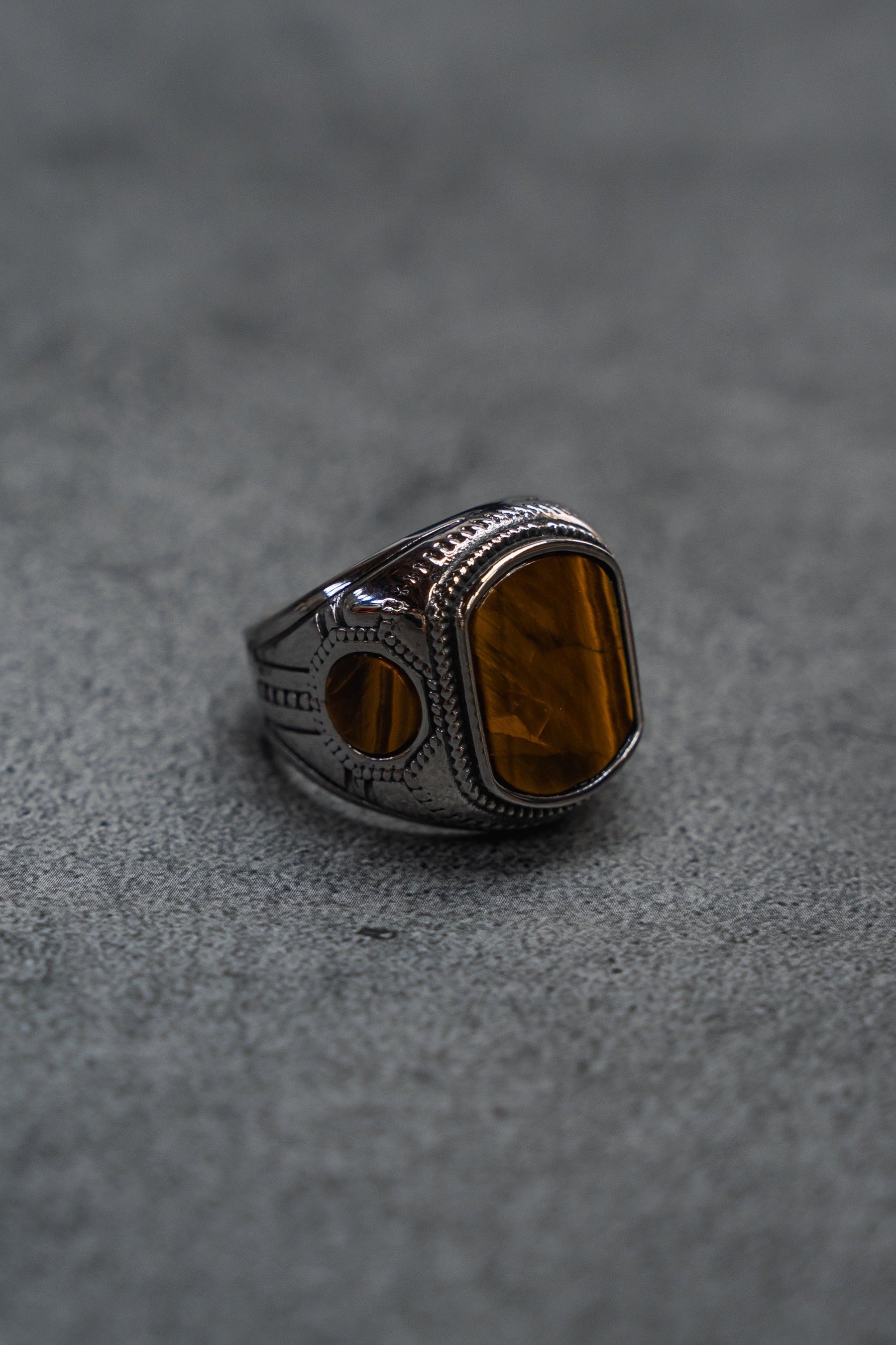 A handcrafted ring from Relentless Betrayal, The Desert Relic Ring, featuring a silver engraved band and a rectangular amber-colored stone, rests on a gray textured surface, capturing its timeless allure.