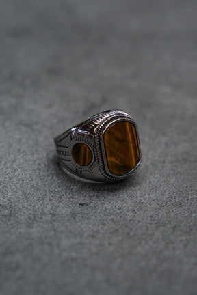 A handcrafted ring from Relentless Betrayal, The Desert Relic Ring, featuring a silver engraved band and a rectangular amber-colored stone, rests on a gray textured surface, capturing its timeless allure.