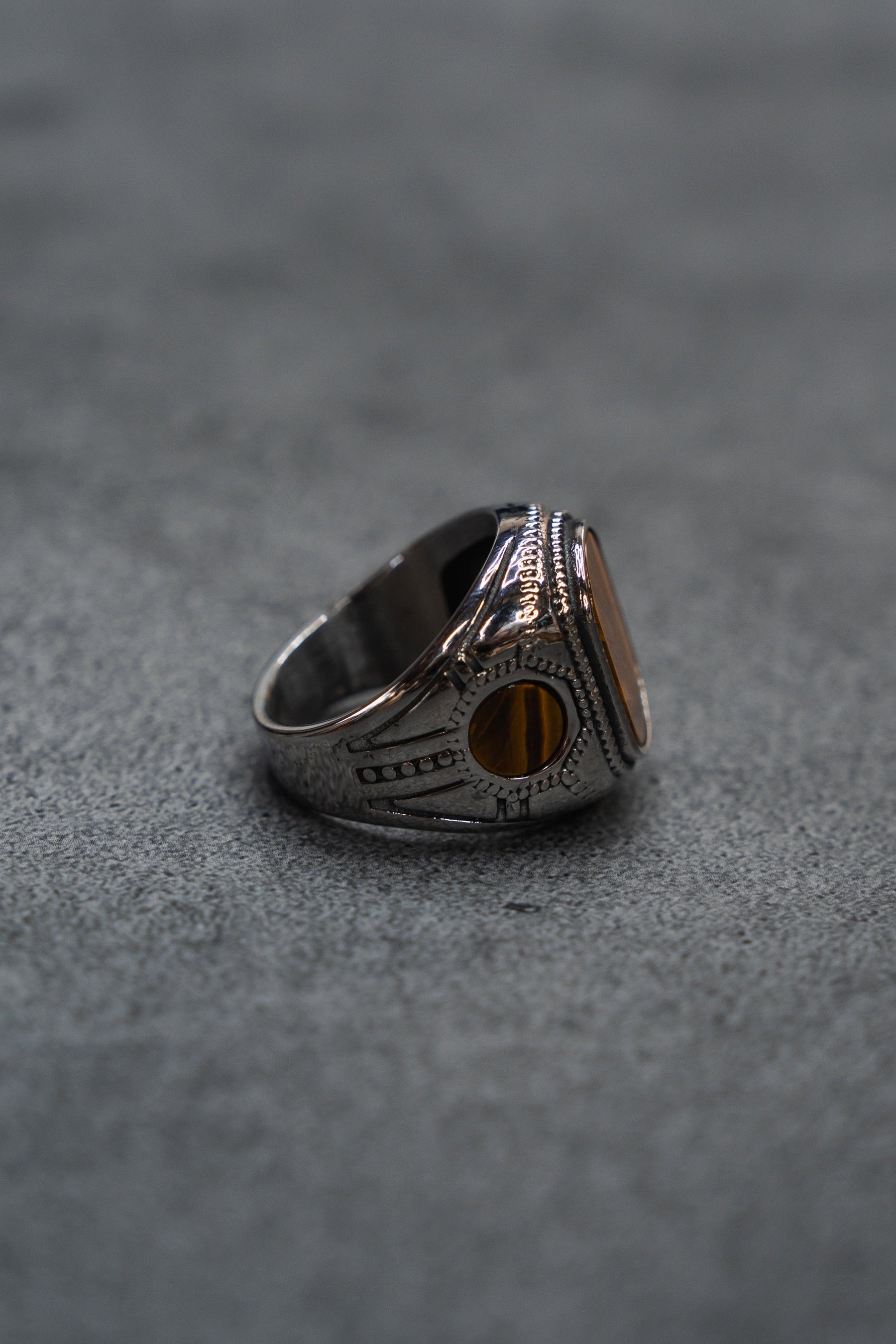 The Desert Relic Ring by Relentless Betrayal, a silver handcrafted piece featuring an oval amber-colored stone, is displayed upright on a gray textured surface and photographed from the side.