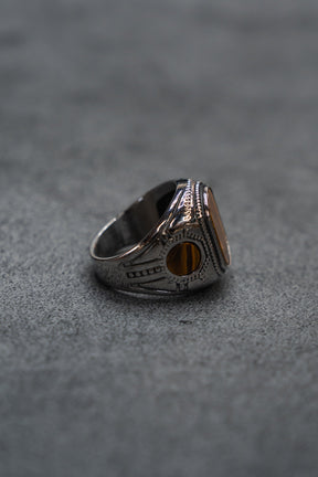 The Desert Relic Ring by Relentless Betrayal, a silver handcrafted piece featuring an oval amber-colored stone, is displayed upright on a gray textured surface and photographed from the side.