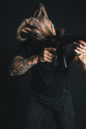 A figure in black clothing dons the Born United Private Label ‘G.O.A.T.’ ram mask and takes aim with a scoped rifle against a dark backdrop—commanding attention with a bold statement, reminiscent of the American tradition found in every iconic patriotic or statement tee.