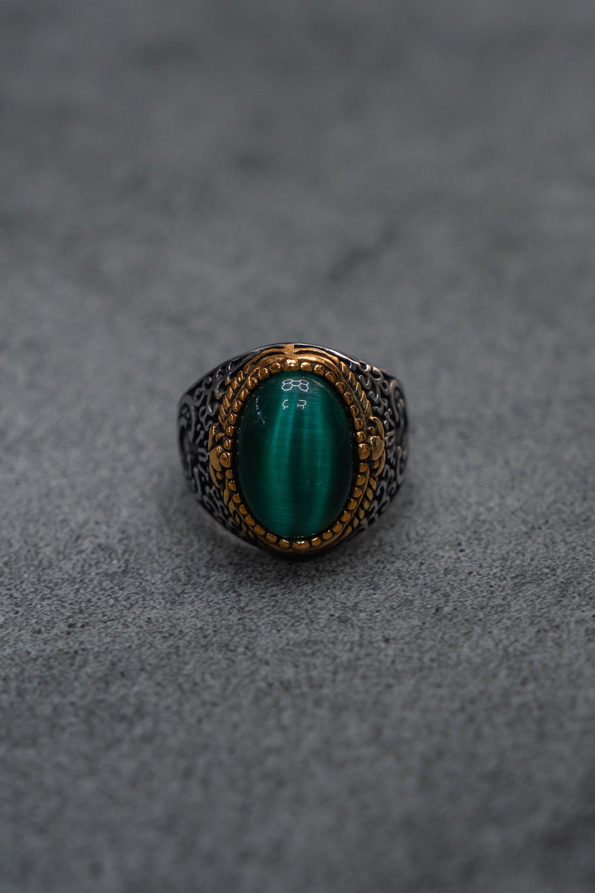 The Relentless Betrayal “Relic Signet Ring” showcases an ornate silver and gold design, featuring a prominent oval green gemstone at its center. The ring is displayed against a subtly weathered gray textured surface.