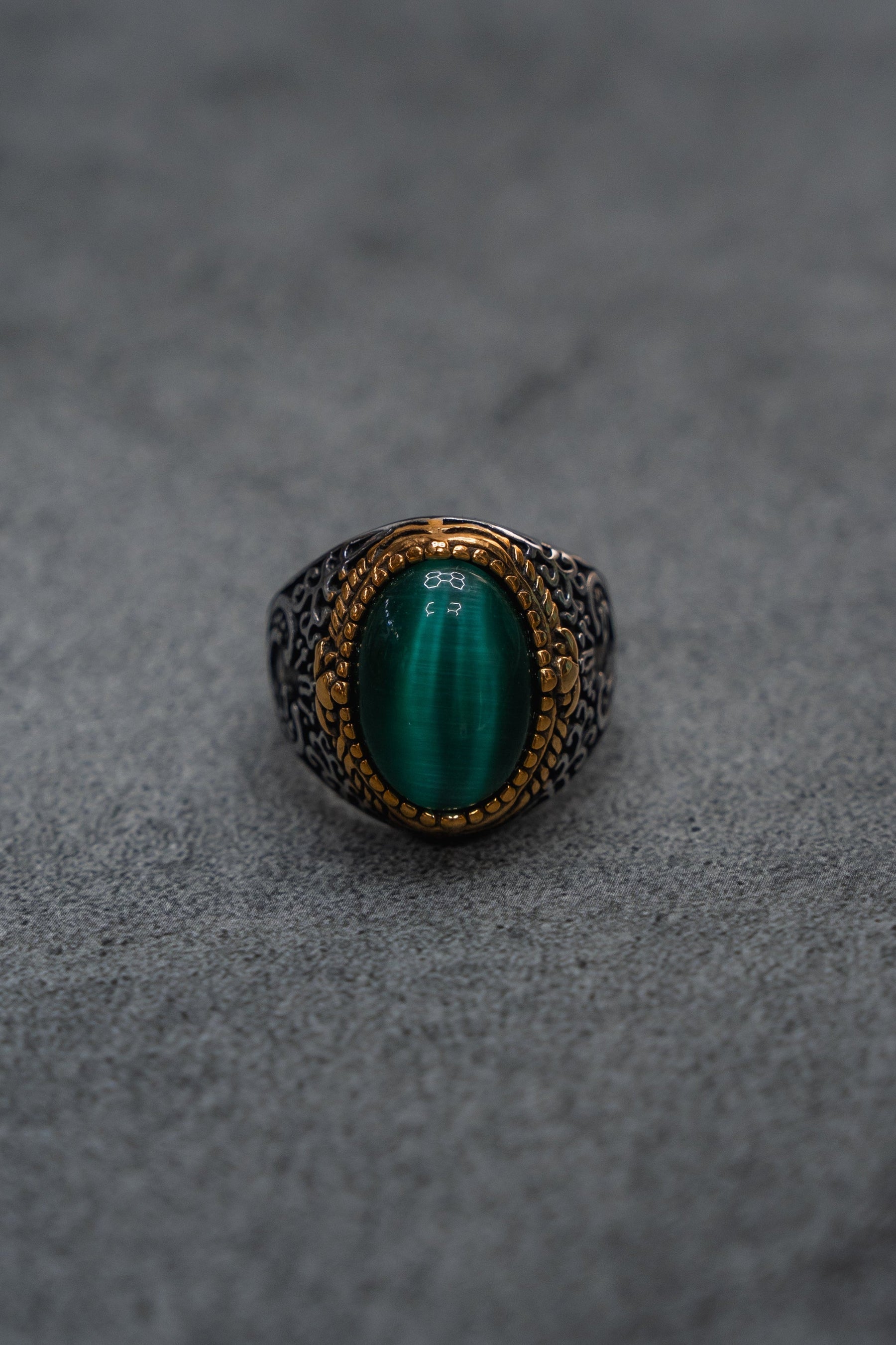 The Relentless Betrayal “Relic Signet Ring” showcases an ornate silver and gold design, featuring a prominent oval green gemstone at its center. The ring is displayed against a subtly weathered gray textured surface.