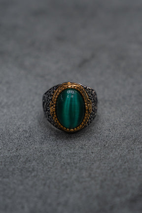 The Relentless Betrayal “Relic Signet Ring” showcases an ornate silver and gold design, featuring a prominent oval green gemstone at its center. The ring is displayed against a subtly weathered gray textured surface.