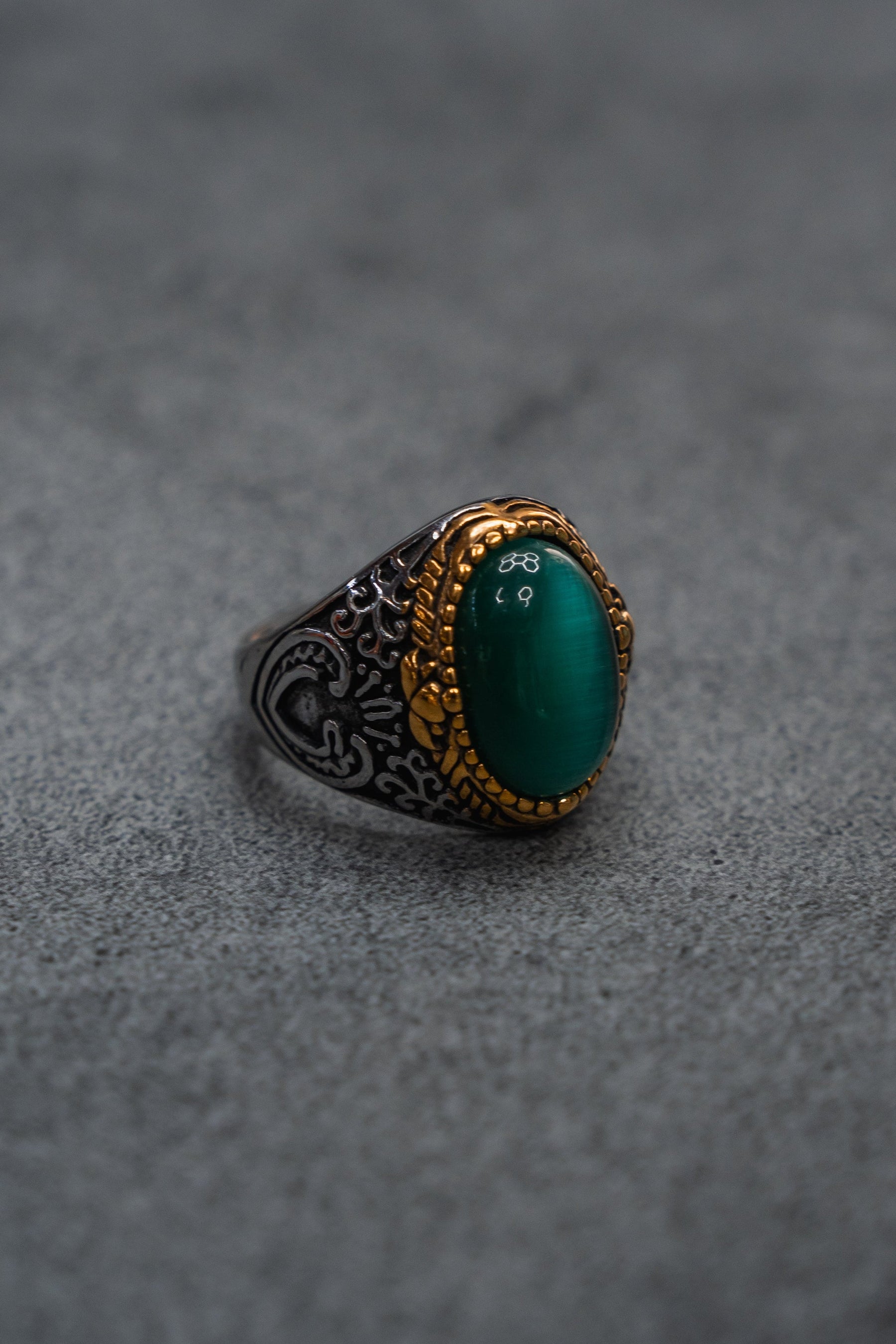 The Relentless Betrayal “Relic Signet Ring” showcases an ornate silver and gold design with a weathered finish, highlighted by an oval deep green stone at its center, and is displayed on a textured gray surface.