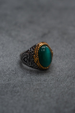 The Relentless Betrayal “Relic Signet Ring” showcases an ornate silver and gold design with a weathered finish, highlighted by an oval deep green stone at its center, and is displayed on a textured gray surface.